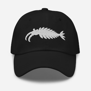 Può includere: Un cappellino da baseball nero con un ricamo bianco raffigurante un Anomalocaris, un artropode marino estinto, centrato sul pannello frontale. Il cappellino presenta una visiera curva, una corona strutturata e un bottone sulla sommità.