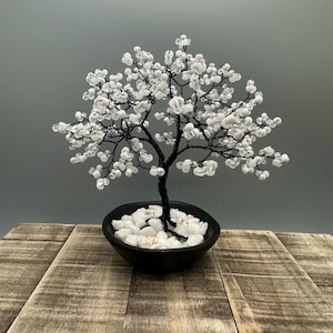 May include: A handcrafted bonsai tree sculpture with a black trunk and branches, adorned with white bead blossoms. The tree sits in a black ceramic bowl filled with white stones, set against a gray backdrop and a wooden surface.