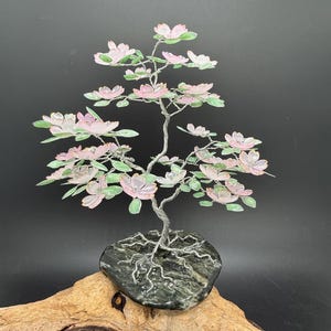 May include: A handcrafted wire tree sculpture with pink and white blossoms and green leaves. The tree is mounted on a dark gray stone base and sits on a piece of driftwood. The tree's branches are made of silver wire.