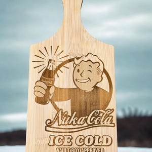 Puede incluir: Tabla de cortar de madera clara con asa. La tabla presenta un diseño grabado con láser del logotipo de Nuka-Cola, un personaje de dibujos animados sosteniendo una botella y las palabras "ICE COLD" y "VAULT BOY APPROVED".