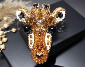 Peut inclure: Broche artisanale en forme de tête de girafe. Elle est ornée de perles dorées, marron, noires et blanches. La girafe a des yeux noirs et de longs cils noirs. La broche est présentée sur un fond sombre, mettant en valeur les détails.