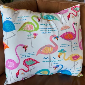 Pu&ograve; includere: Un cuscino quadrato con un motivo di fenicotteri colorati su sfondo bianco. I fenicotteri, con occhiali da sole e cappelli, sono raffigurati in varie pose, con onde e gelati. Il cuscino &egrave; in tessuto e si trova su una superficie marrone.