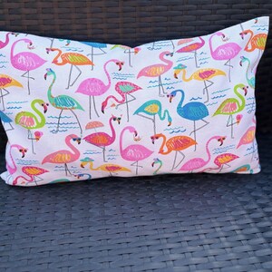 Pu&ograve; includere: Cuscino rettangolare decorativo con sfondo bianco e motivo di fenicotteri colorati. I fenicotteri sono in vari colori, tra cui rosa, arancione, giallo, verde e blu. Il cuscino &egrave; appoggiato su una sedia di vimini marrone scuro.