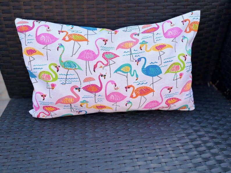 Pu&ograve; includere: Cuscino rettangolare decorativo con sfondo bianco e motivo di fenicotteri colorati. I fenicotteri sono in vari colori, tra cui rosa, arancione, giallo, verde e blu. Il cuscino &egrave; appoggiato su una sedia di vimini marrone scuro.