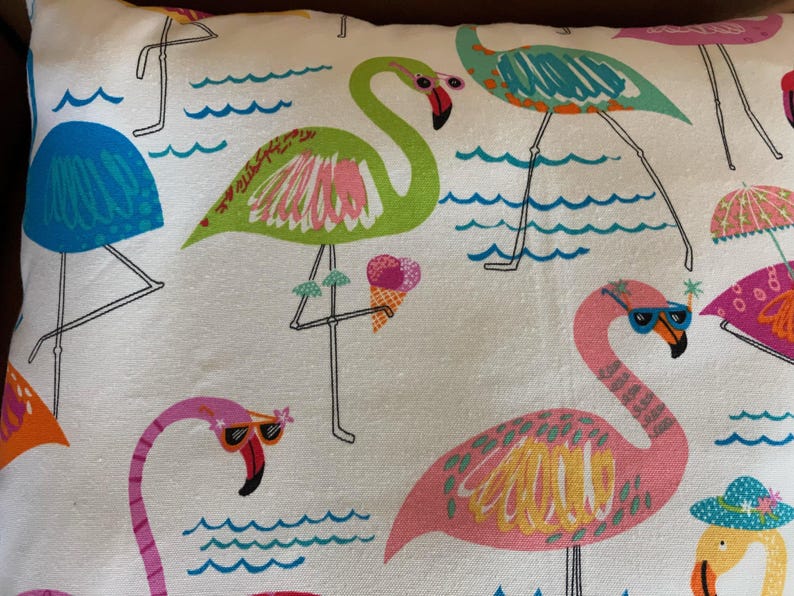 Pu&ograve; includere: Cuscino bianco con un motivo colorato di fenicotteri. Il design presenta vari fenicotteri nei toni del rosa, verde e blu, alcuni con occhiali da sole e gelati. Lo sfondo include acqua stilizzata e ombrelloni.