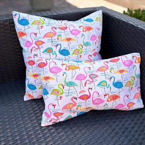 Pu&ograve; includere: Due cuscini decorativi con sfondo bianco e un colorato motivo a fenicotteri. I fenicotteri sono in tonalit&agrave; di rosa, arancione, blu e verde. I cuscini sono rettangolari e quadrati, appoggiati su una sedia in vimini marrone scuro.