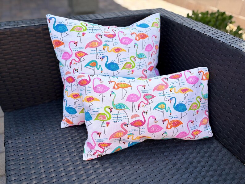 Pu&ograve; includere: Due cuscini decorativi con sfondo bianco e un colorato motivo a fenicotteri. I fenicotteri sono in tonalit&agrave; di rosa, arancione, blu e verde. I cuscini sono rettangolari e quadrati, appoggiati su una sedia in vimini marrone scuro.