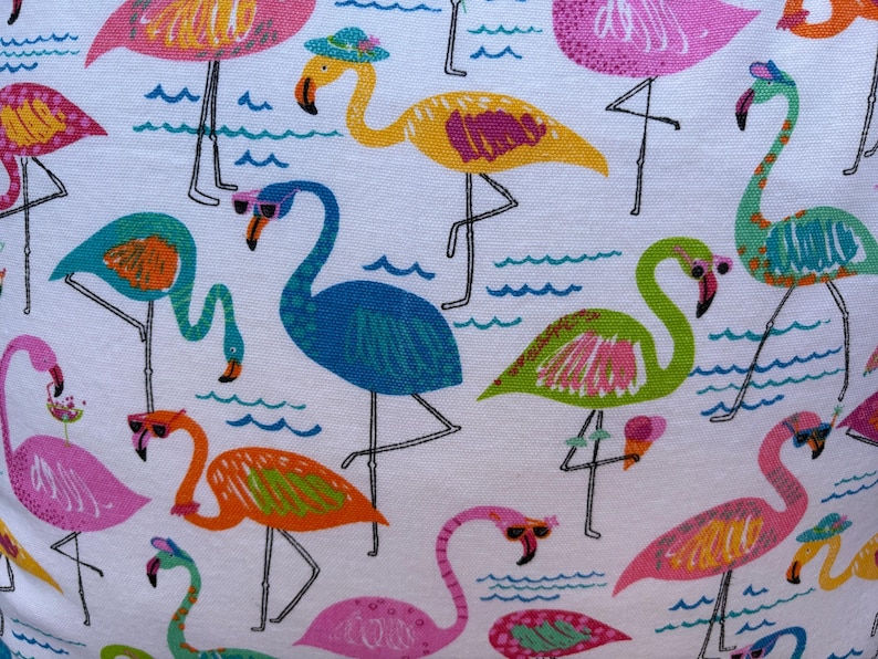 Pu&ograve; includere: Tessuto bianco con un motivo ripetuto di colorati fenicotteri. I fenicotteri sono raffigurati in rosa, blu, verde e arancione, alcuni con occhiali da sole o cappelli. Linee ondulate blu rappresentano l'acqua.