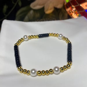 May include: A beaded bracelet featuring a repeating pattern of small metallic gold spheres, smooth white pearls, and cylindrical black beads. The circular accessory is arranged with alternating gold and black segments, accented by four evenly spaced white pearls.
