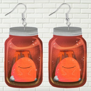 May include: A pair of red glass jar earrings with a small orange creature inside. The creature is sitting on a small volcano with a brown and gray rock base. The jars have silver lids and silver earring hooks.
