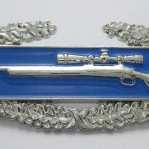 Puede incluir: Una insignia de experto en francotirador del ejército de plata con un centro de esmalte azul que presenta un rifle de plata con un visor. La insignia está rodeada por una corona de plata de hojas de roble.