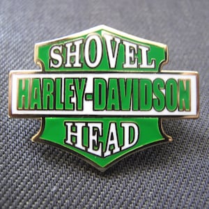 Puede incluir: Un pin metálico verde y dorado con las palabras "SHOVEL HARLEY-DAVIDSON HEAD" en letras blancas. El pin tiene forma rectangular con borde dorado. El fondo es de tela gris oscuro texturizada. Diseño clásico.