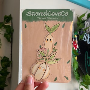 May include: A sticker sheet featuring two cartoon turnip characters with smiling faces and green leaves. The sticker sheet is packaged in a clear plastic sleeve and has the text "Sacred Cove Co. Turnip Reunion" printed on it.
