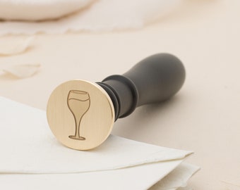 May include: A gold-toned metal stamp with a black handle. The stamp features a design of a wine glass.