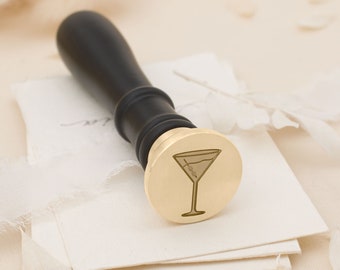 May include: A gold-toned wax seal stamp with a martini glass design. The stamp is attached to a black wooden handle.
