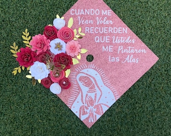 Op de afbeelding: Een roze afstudeerhoed versierd met papieren bloemen en gouden accenten. De hoed heeft een wit beeld van de Maagd Maria en de tekst "CUANDO ME Vean Volar RECUERDEN QUE Ustedes ME Pintaron las Alas".