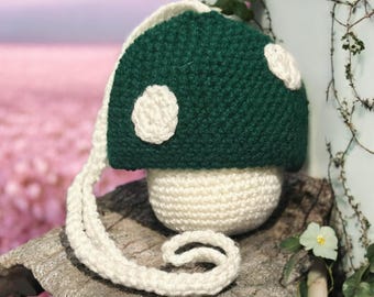 May include: A handmade crochet mushroom bag in green and white. The mushroom cap is dark green with white spots, and the stem is white. The bag has a long white strap and is sitting on a piece of wood.