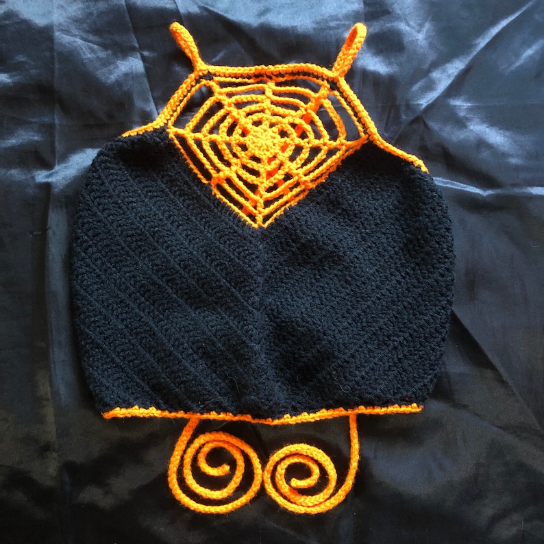 May include: A black crochet top with an orange crocheted spiderweb design on the front. The top has a halter neck style and a tie closure at the back.