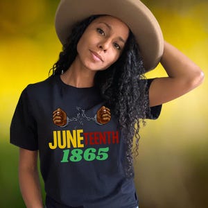 May include: A woman wearing a black t-shirt with the text "Juneteenth 1865" and two raised fists in red, brown, and yellow colours. The woman is wearing a brown hat and has curly black hair.