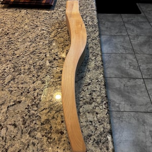 May include: A long, curved piece of wood with a natural finish. The wood is light brown in color and has a smooth surface. The wood is lying on a countertop.