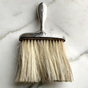 Puede incluir: Un cepillo de tocador plateado con mango y cabezal rectangular ancho. El mango y el marco del cabezal del cepillo presentan grabados florales. Las cerdas son de color beige claro, sobre un fondo de mármol.