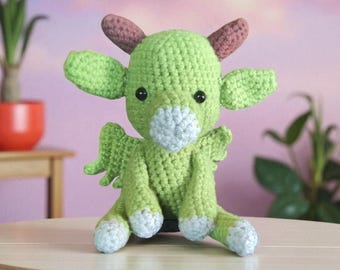Puede incluir: Un dragón de ganchillo hecho a mano en tonos verdes, con pequeñas alas, orejas y cuernos. El dragón tiene ojos negros y un hocico y pies azul claro. El peluche está sentado sobre una superficie de madera.