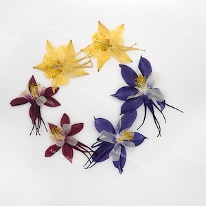 May include: A circular arrangement of pressed flowers in shades of yellow, deep purple, and burgundy. The delicate petals and long stamens are visible. The flowers are arranged on a white background. The text "AvisFloralsAndGifts" is visible.