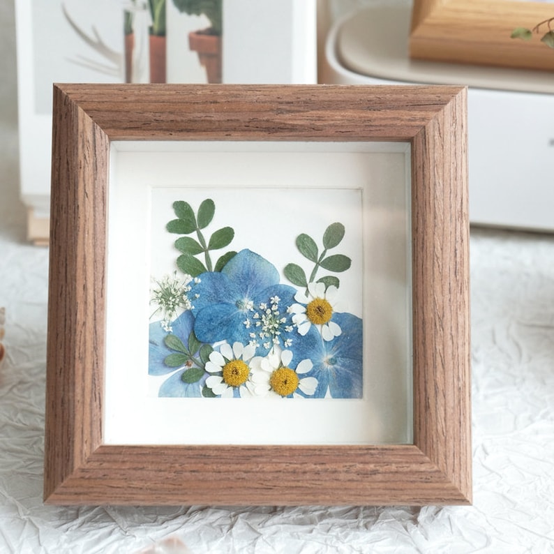May include: A framed pressed flower art piece with blue and white flowers and green leaves. The frame is made of dark wood.