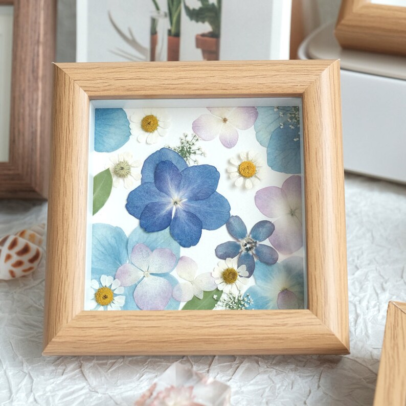 May include: A framed pressed flower art piece with a white background and a variety of blue, purple, and white flowers. The frame is made of light wood.