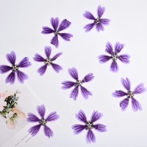May include: A collection of eight dried purple flowers arranged in a symmetrical pattern on a white background.