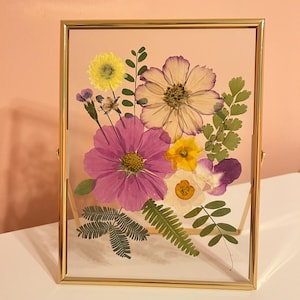 May include: A framed floral arrangement featuring pressed flowers in shades of purple, yellow, and white. The flowers are arranged on a light pink background within a gold-colored metal frame. The frame is rectangular and has a clear glass front.