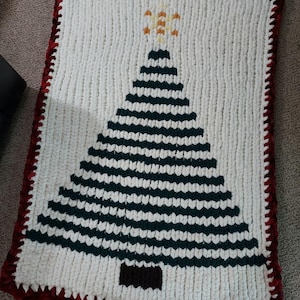 May include: A rectangular crochet blanket featuring a white background with a large, centered dark green triangular Christmas tree design. The tree is topped with a yellow star and has a brown trunk. The blanket has a dark red border.