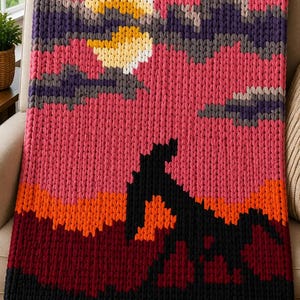 May include: A chunky knit throw blanket featuring a landscape design with a pink sky, yellow moon, and dark purple clouds. The lower section depicts black and dark red mountains with orange highlights, finished with a row of black fringe at the bottom.