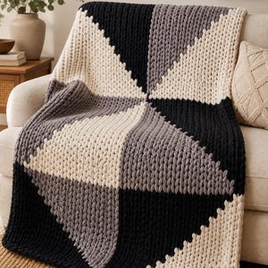 May include: A chunky knit throw blanket featuring a geometric pattern of large triangles in alternating colors of black, cream, and charcoal gray. The blanket is constructed with thick, soft yarn in a prominent braided stitch texture throughout.
