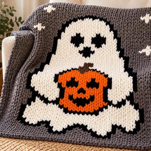 May include: A chunky knit throw blanket in dark gray featuring a large white ghost character holding a bright orange jack-o'-lantern pumpkin. The design includes small white star accents against the gray background, crafted with thick yarn.