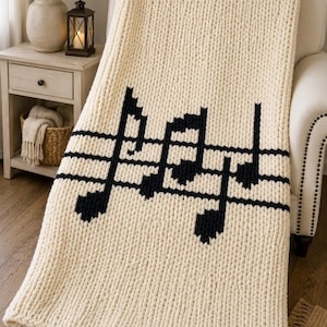 May include: A chunky knit throw blanket in cream white featuring a large black musical staff pattern with various musical notes. The blanket has a thick, textured yarn construction and is draped over a piece of furniture, displaying a clear contrast.