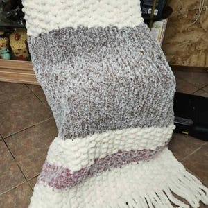 May include: A chunky knit throw blanket featuring horizontal stripes in cream, mauve, and a variegated grey and white yarn. The blanket is constructed with thick, soft yarn loops and is finished with long, cream-colored fringe along the bottom edge.