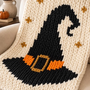 May include: A chunky knit throw blanket in cream featuring a large black witch hat design with an orange band and a gold buckle. The blanket is decorated with small gold star accents scattered across the cream background in a thick yarn texture.