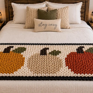 Könnte beinhalten: Ein cremefarbener, grob gestrickter Bettläufer mit drei großen Kürbismotiven in Orange, Beige und Rostrot. Jeder Kürbis hat einen dunkelbraunen Stiel und ein grünes Blatt. Der Läufer ist mit einem dicken, dunkelbraunen geflochtenen Rand versehen.