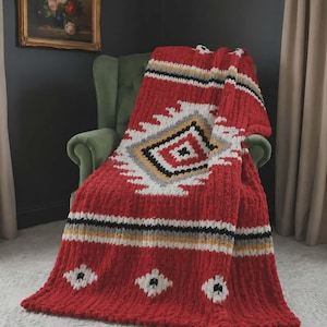 May include: A chunky knit throw blanket featuring a vibrant red base with a geometric pattern in white, black, and yellow. The design includes a central diamond motif and horizontal stripes, crafted from thick, soft yarn with a textured finish.