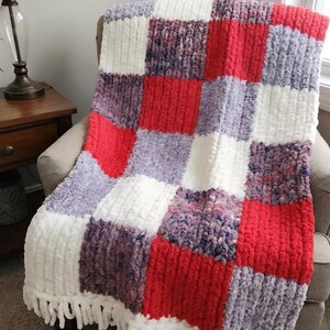 May include: A handmade, plush blanket in a patchwork design. The blanket features squares of red, white, and shades of purple. It has a white fringe along the bottom edge. The blanket is draped over a chair.