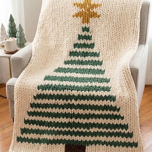 May include: A chunky knit throw blanket in cream yarn featuring a large, centered Christmas tree design in dark green with a mustard yellow star at the top and a brown trunk at the base, draped over a gray armchair with a small leather brand tag.