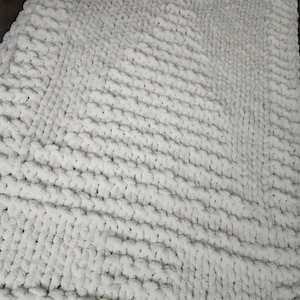 May include: A rectangular white blanket crafted from thick, chunky yarn with a prominent knit texture. The design features a central panel with a subtle, raised pattern resembling a tree shape, surrounded by a wide, textured border.