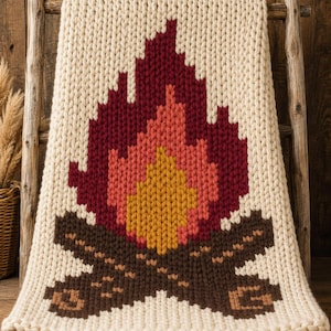 May include: A chunky knit blanket in cream yarn featuring a large, centered graphic of a campfire. The fire design is composed of deep burgundy, bright coral, and golden yellow stitches, while the logs are depicted in dark brown and tan yarn.