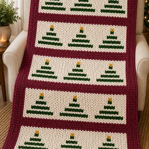 Könnte beinhalten: Eine grob gestrickte Decke mit einem tief burgunderroten Rand und fünf horizontalen cremefarbenen Bahnen. Jede Bahn enthält drei grüne, dreieckige Baummotive mit kleinen gelben Sternen an der Spitze, die in einem wiederkehrenden Muster angeordnet sind.