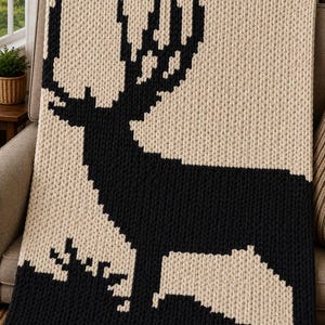 May include: A chunky knit throw blanket featuring a large, black, pixelated silhouette of a deer with prominent antlers against a cream-colored background. The blanket is constructed from thick, soft yarn with a visible, textured stitch pattern.