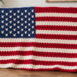Puede incluir: Una manta de punto grueso con el diseño de la bandera estadounidense. La manta está confeccionada con hilo grueso en colores azul profundo, rojo brillante y blanco roto, con pequeñas puntadas blancas en forma de V que forman las estrellas.