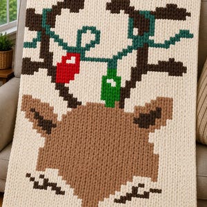 May include: A cream-colored chunky knit blanket featuring a large brown reindeer face with dark brown antlers. The antlers are decorated with a green string of lights and two prominent light bulbs in solid red and bright green colors.
