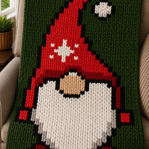 May include: A chunky knit blanket featuring a large gnome graphic. The gnome has a tall red hat with a white star pattern, a beige nose, and a long white beard. The background of the blanket is dark green, and the gnome is outlined in black.