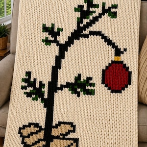Puede incluir: Una manta de punto grueso color crema con un diseño pixelado de un árbol negro escaso en una maceta color canela. El árbol tiene pequeñas hojas verdes y un solo adorno navideño rojo grande que cuelga de una rama curva de lana gruesa.